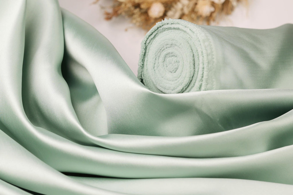 A close-up view of a light green satin fabric, possibly silk, with a rolled-up end visible.