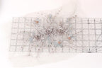 a decorative floral arrangement with three large white flowers and smaller silver flowers, all adorned with small beads. The arrangement is displayed on a transparent ruler, which is placed on a white surface.