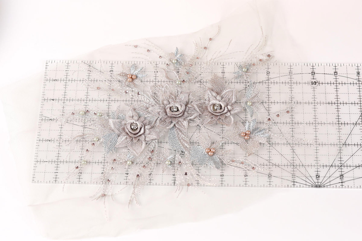 a decorative floral arrangement with three large white flowers and smaller silver flowers, all adorned with small beads. The arrangement is displayed on a transparent ruler, which is placed on a white surface.