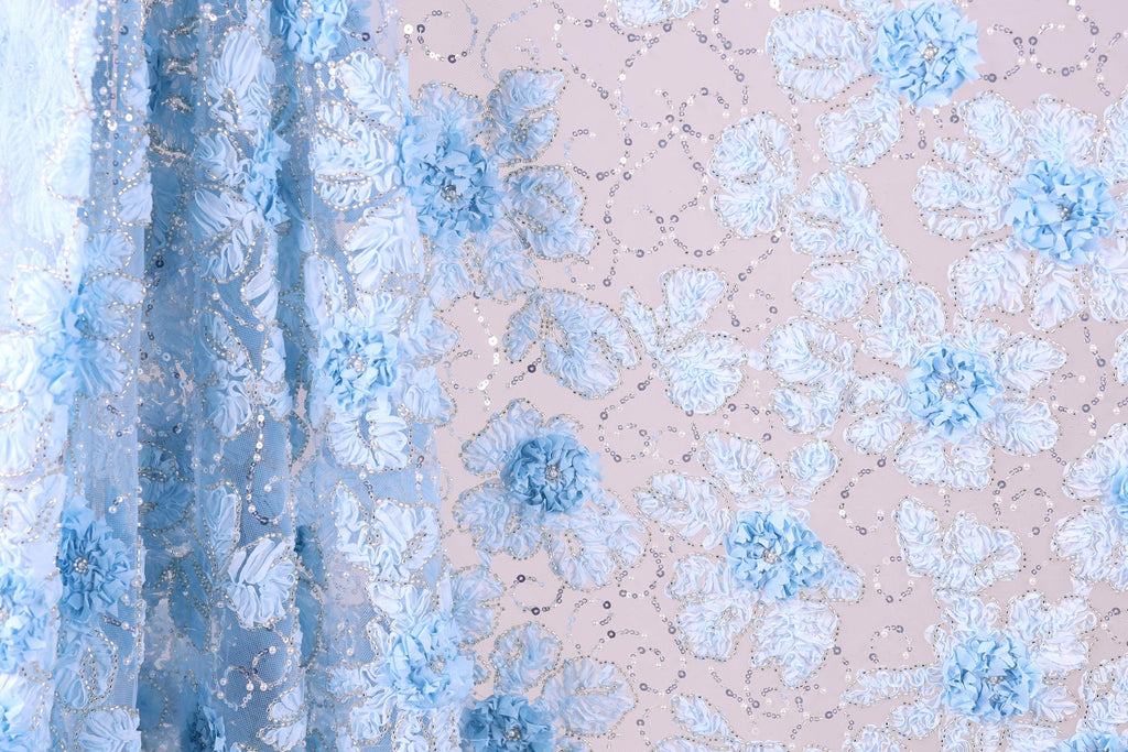 a close-up view of a fabric with a floral pattern, predominantly in shades of blue and white. The fabric appears to be made of a sheer material, with a delicate and intricate design of flowers and leaves. The background of the image is a light pink color, which complements the blue and white floral pattern on the fabric.