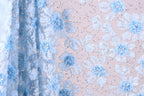 a close-up view of a fabric with a floral pattern, predominantly in shades of blue and white. The fabric appears to be made of a sheer material, with a delicate and intricate design of flowers and leaves. The background of the image is a light pink color, which complements the blue and white floral pattern on the fabric.