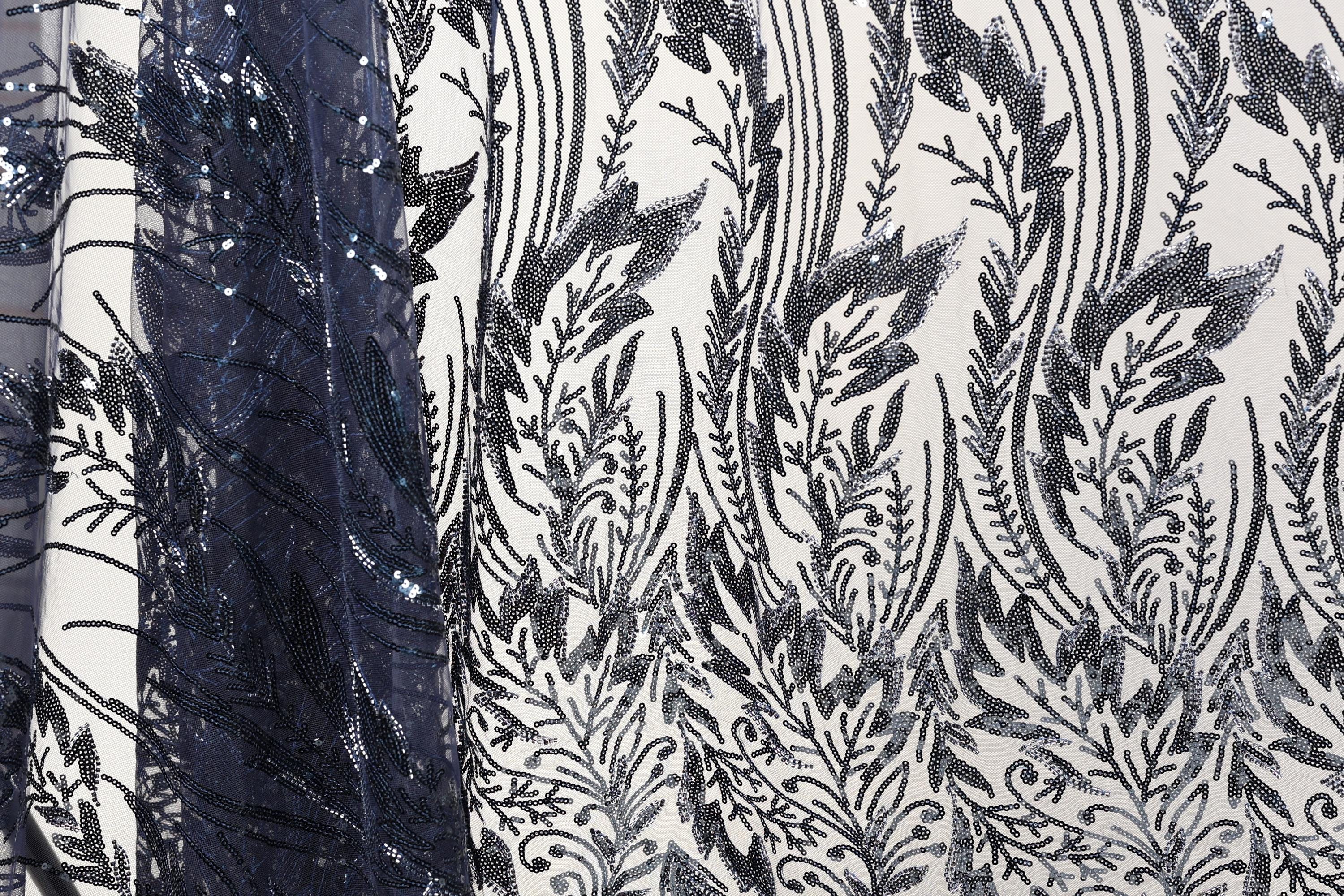 a close-up view of a fabric with a black and white floral pattern. The fabric appears to be made of a material with sequins or beads, adding a decorative element to the design. The pattern is intricate and detailed, showcasing the craftsmanship and artistry involved in creating the fabric.
