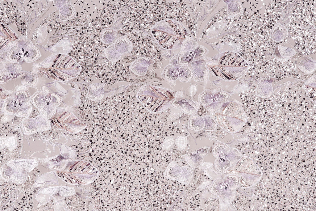 a close-up view of a fabric with a floral pattern, including white and purple flowers and sequins. The fabric appears to be a light-colored material, possibly lace or a similar type of textile. The pattern is intricate and detailed, with the flowers and sequins adding texture and visual interest to the fabric.