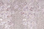 a close-up view of a fabric with a floral pattern, including white and purple flowers and sequins. The fabric appears to be a light-colored material, possibly lace or a similar type of textile. The pattern is intricate and detailed, with the flowers and sequins adding texture and visual interest to the fabric.