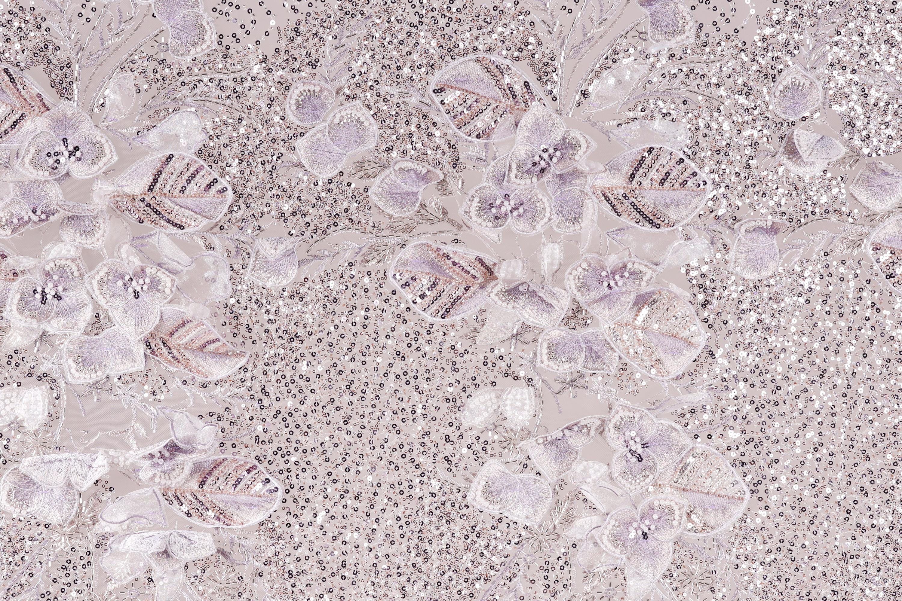 a close-up view of a fabric with a floral pattern, including white and purple flowers and sequins. The fabric appears to be a light-colored material, possibly lace or a similar type of textile. The pattern is intricate and detailed, with the flowers and sequins adding texture and visual interest to the fabric.