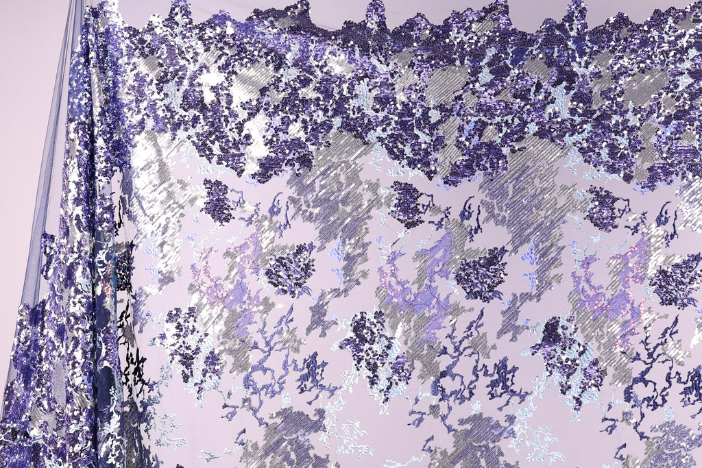 a fabric with a purple and white pattern, with a mix of small and large sequins or beads scattered across the fabric. The pattern appears to be a blend of different shades of purple and white, creating a visually striking and intricate design.