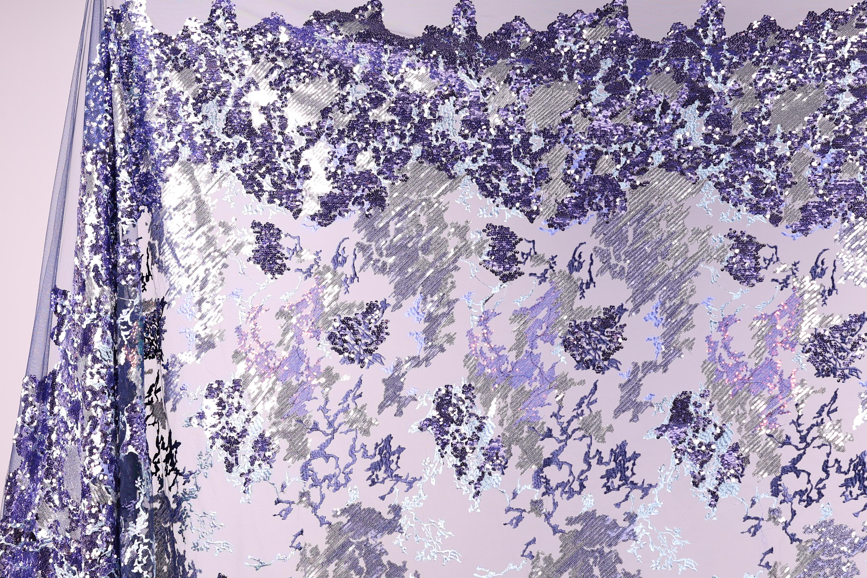 a fabric with a purple and white pattern, with a mix of small and large sequins or beads scattered across the fabric. The pattern appears to be a blend of different shades of purple and white, creating a visually striking and intricate design.