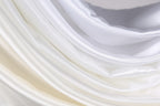 The image is a close-up of a soft, flowing fabric with a gradient of white and beige colors. The fabric appears to be made of silk or satin, and it has a smooth, shiny texture. The colors are predominantly white and beige, with some variations in the shades of the fabric. The fabric is folded and draped in a way that creates a sense of movement and softness.