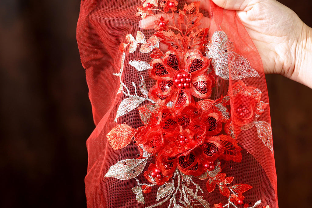 A hand is holding a red fabric with a floral design, including red flowers and leaves, as well as red sequins.