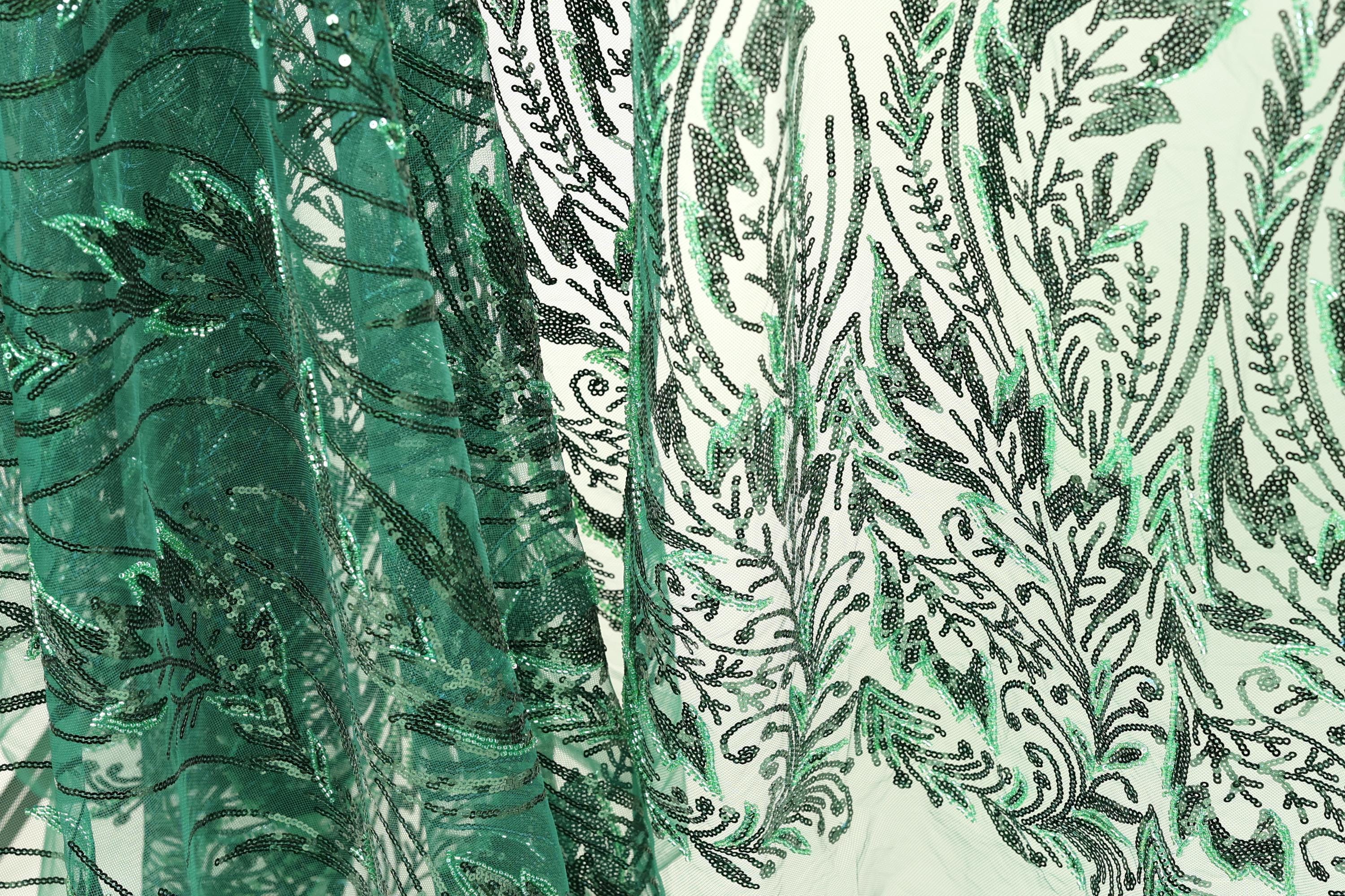 a close-up view of a fabric with a green and black floral pattern. The fabric appears to be made of a material with a textured surface, possibly lace or a similar type of material. The pattern consists of various shapes and designs, including leaves, flowers, and other decorative elements, creating a visually striking and intricate design.