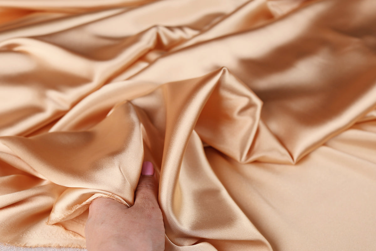 A hand is holding a piece of light brown satin fabric, which appears to be soft and smooth.