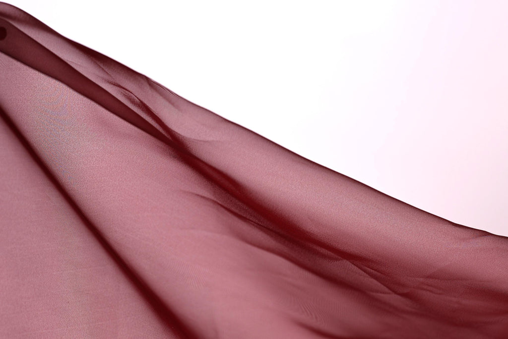 A close-up view of a flowing, maroon-colored fabric, possibly a sheer or sheer-like material, against a plain white background.