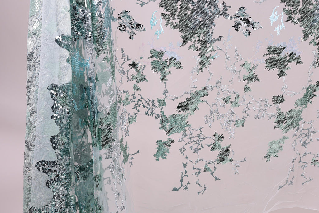 a close-up view of a fabric with a floral pattern, including green and white elements. The fabric appears to be made of a sheer material, with a metallic or sequined texture. The pattern is intricate and detailed, creating a visually appealing and textured appearance.