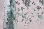 a close-up view of a fabric with a floral pattern, including green and white elements. The fabric appears to be made of a sheer material, with a metallic or sequined texture. The pattern is intricate and detailed, creating a visually appealing and textured appearance.