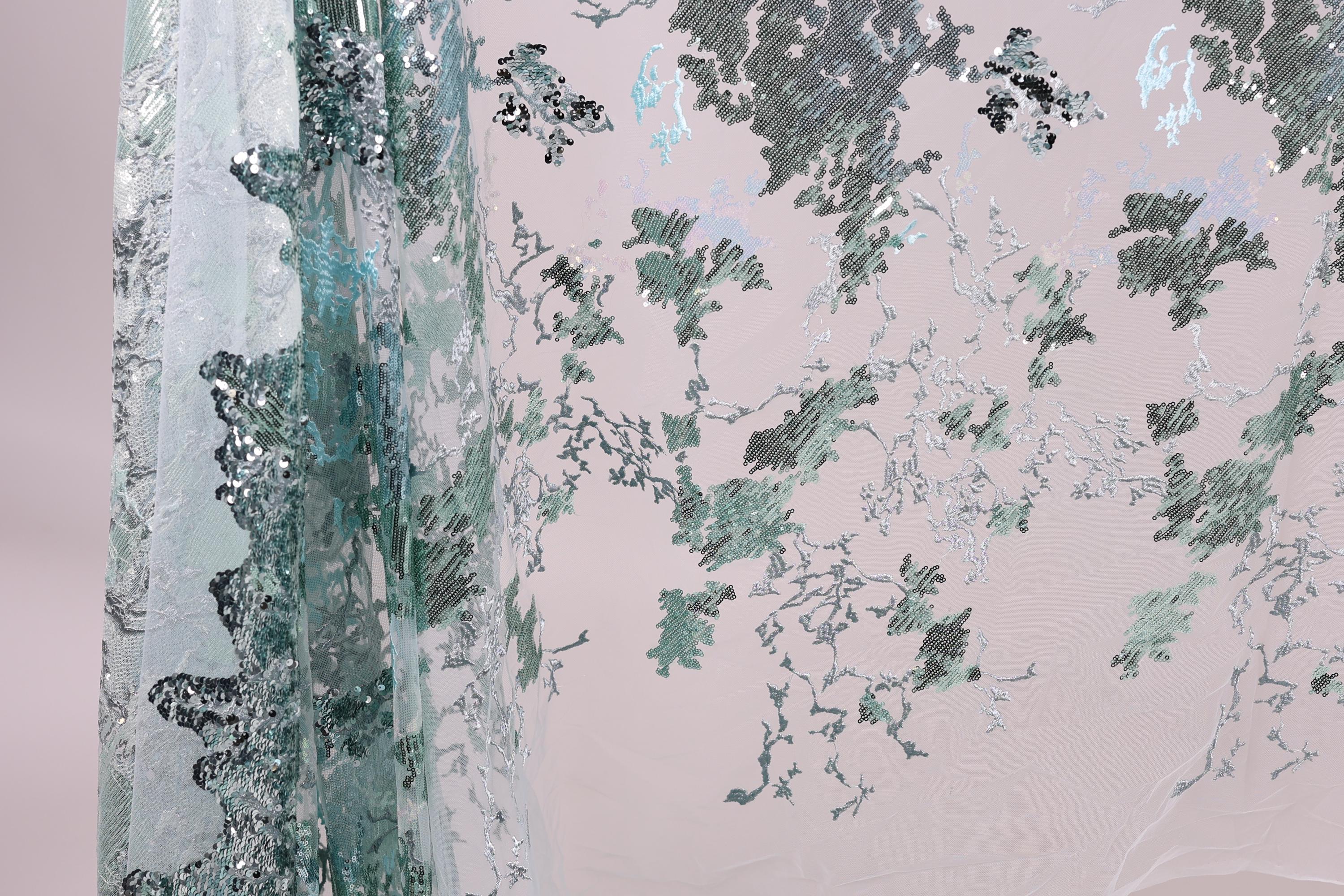 a close-up view of a fabric with a floral pattern, including green and white elements. The fabric appears to be made of a sheer material, with a metallic or sequined texture. The pattern is intricate and detailed, creating a visually appealing and textured appearance.