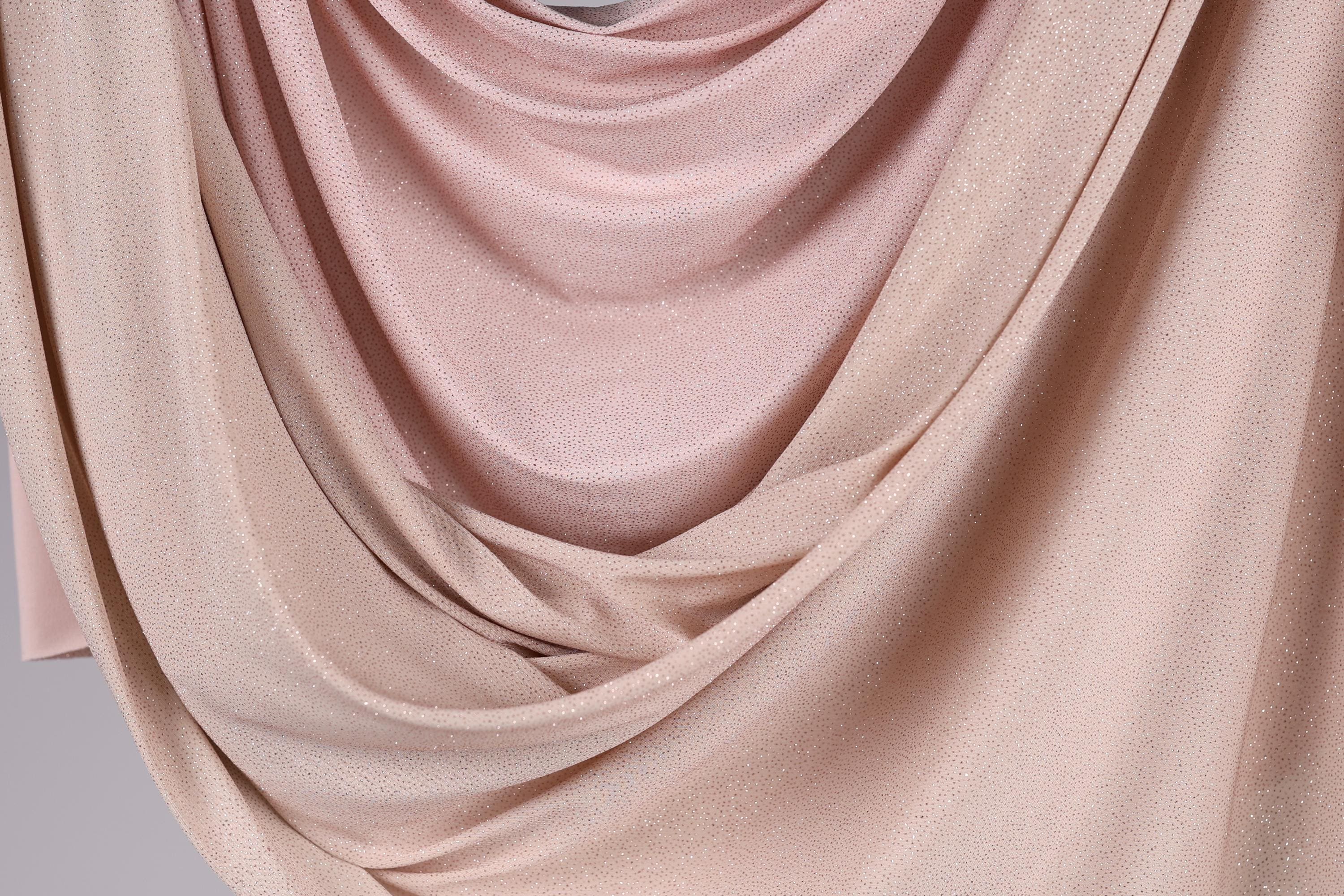 a close-up view of a fabric with a soft, flowing texture in shades of pink and beige. The fabric appears to be made of a shiny, glittery material, giving it a delicate and elegant appearance.