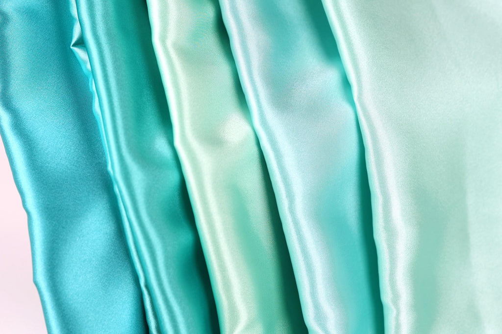 A close-up image of a collection of turquoise-colored satin fabric, with varying shades of turquoise visible.