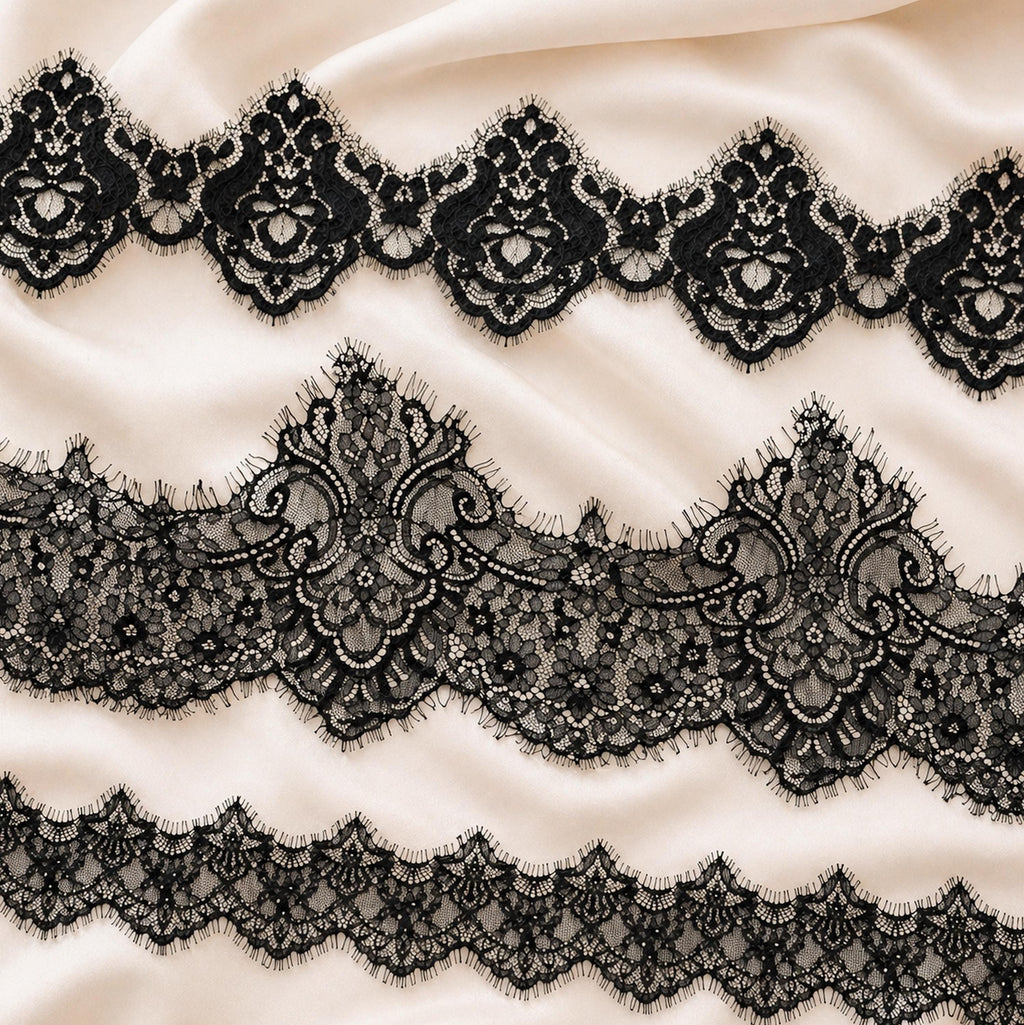 a close-up view of a delicate, intricate black lace pattern on a soft, light-colored fabric. The lace appears to be made of silk and is adorned with ornate, floral-inspired designs. The fabric has a smooth, flowing texture, and the lace is arranged in a repeating pattern across the entire surface.