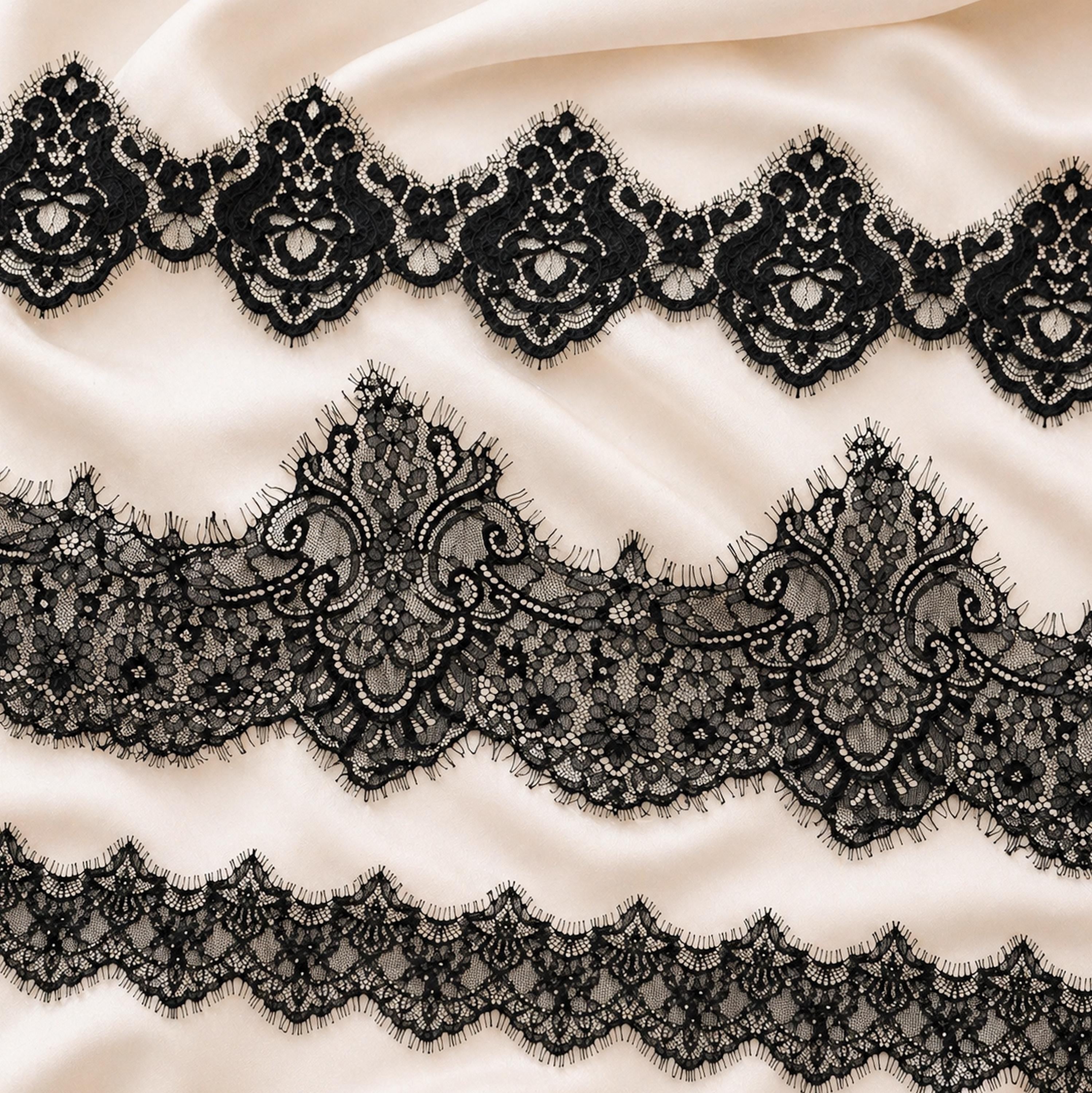 a close-up view of a delicate, intricate black lace pattern on a soft, light-colored fabric. The lace appears to be made of silk and is adorned with ornate, floral-inspired designs. The fabric has a smooth, flowing texture, and the lace is arranged in a repeating pattern across the entire surface.