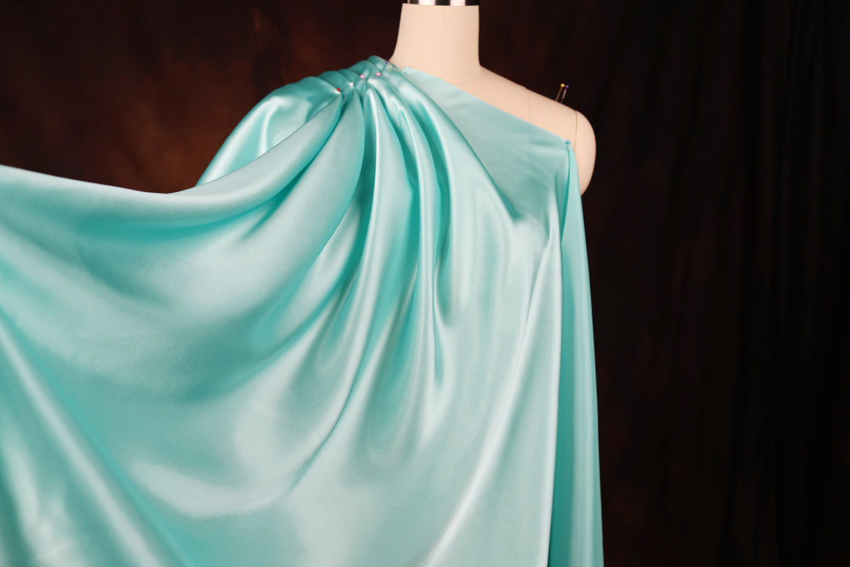 A mannequin wearing a flowing, turquoise satin dress with a draped, one-shoulder design.