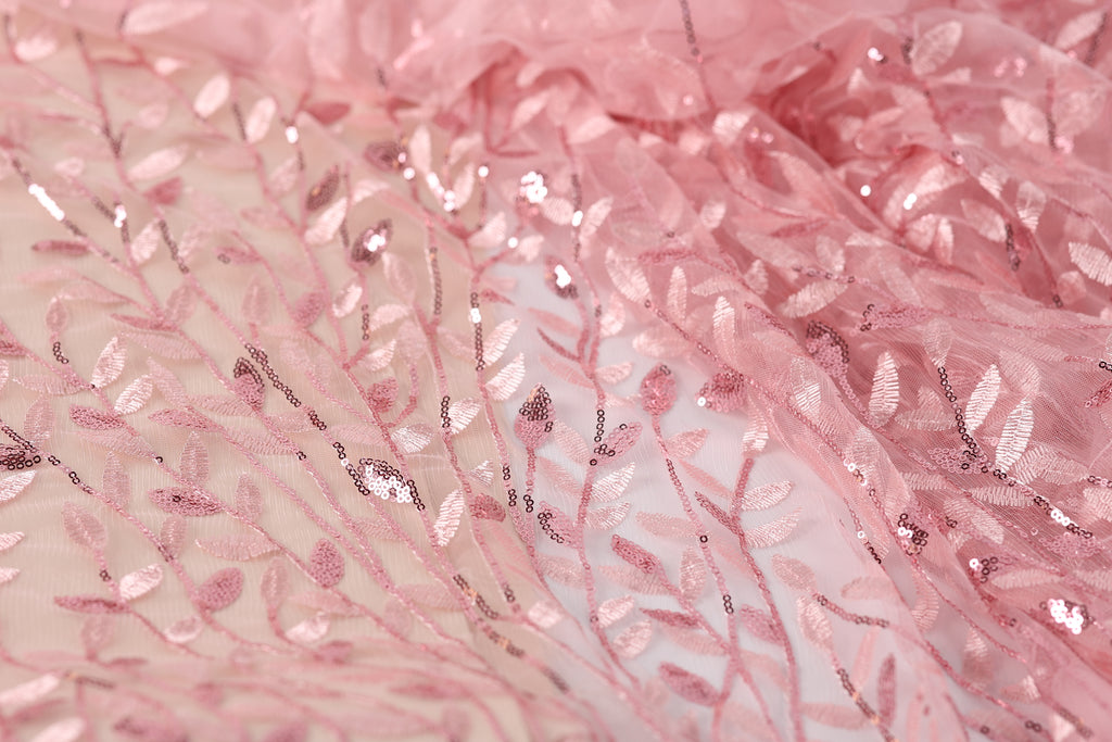 a close-up view of a delicate, pink fabric with a floral pattern. The fabric appears to be made of sheer material, with a lace-like texture and a shimmering, sequined effect. The pattern consists of small, repeating leaves and flowers, creating a delicate and intricate design. The fabric is the main focus of the image, with no other objects or text present.