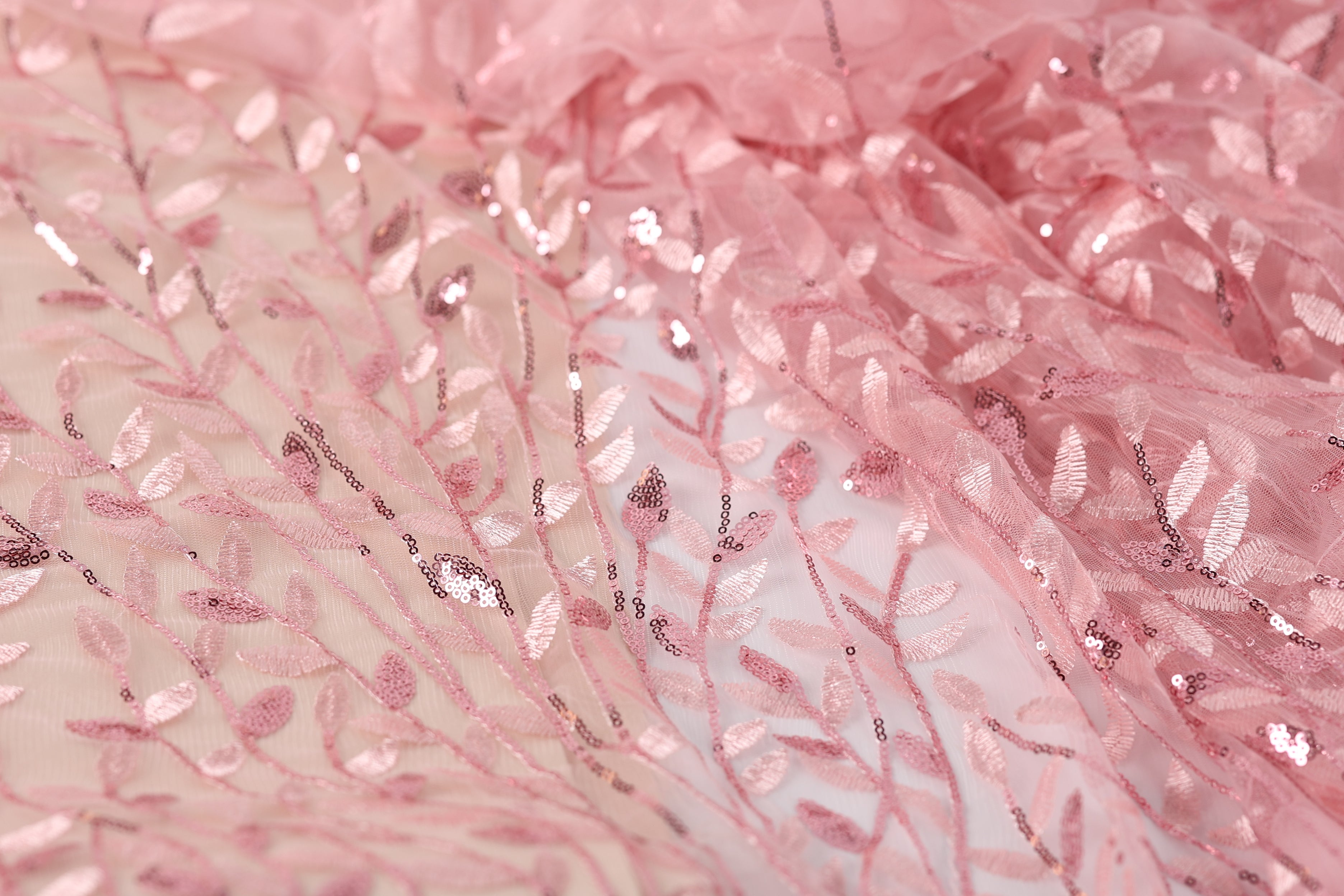 a close-up view of a delicate, pink fabric with a floral pattern. The fabric appears to be made of sheer material, with a lace-like texture and a shimmering, sequined effect. The pattern consists of small, repeating leaves and flowers, creating a delicate and intricate design. The fabric is the main focus of the image, with no other objects or text present.