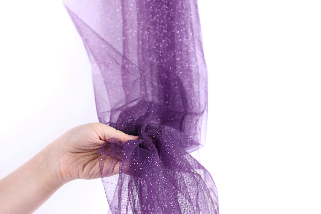A hand is holding a piece of purple glittery fabric, which appears to be a sheer or sheer-like material.