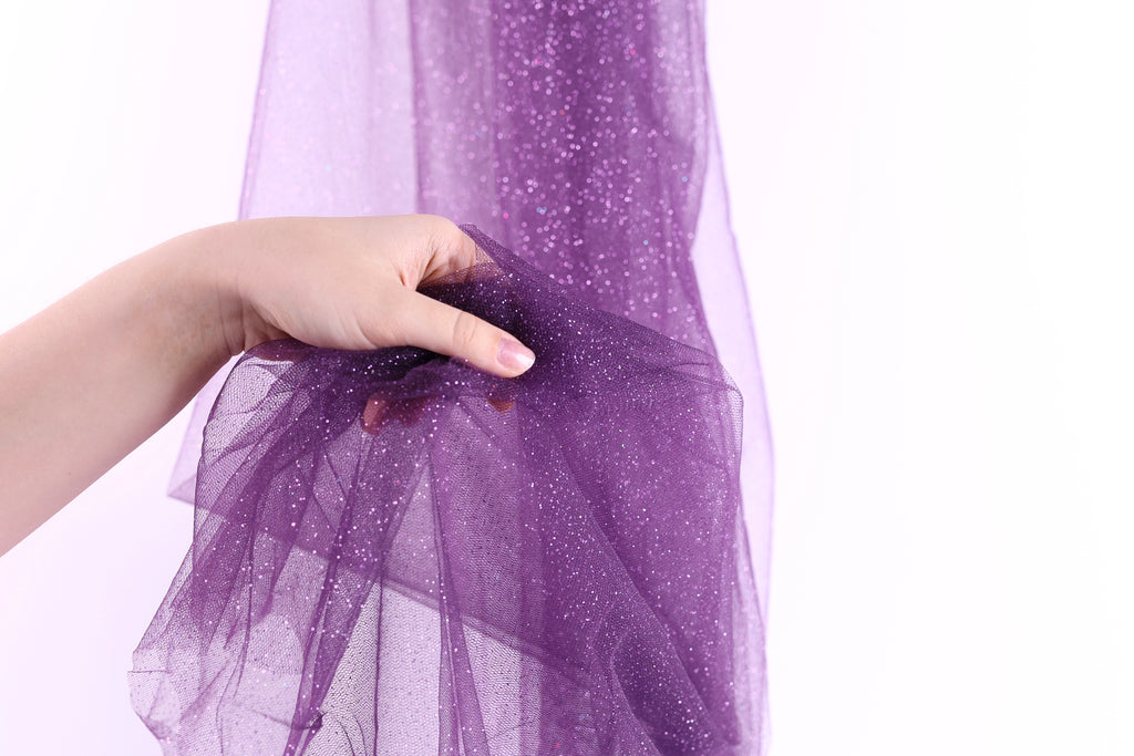 A hand is holding a piece of purple glittery fabric, which appears to be a sheer or sheer-like material.