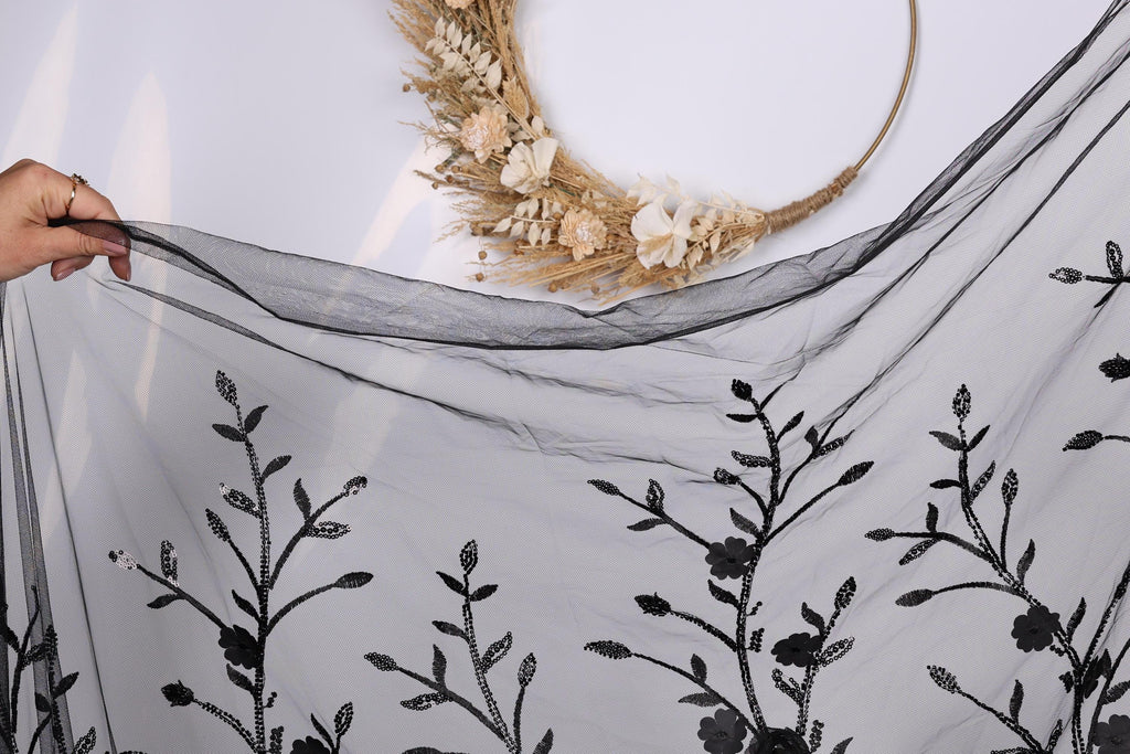 a hand holding a black fabric with a floral pattern, including leaves and flowers, against a white background.
