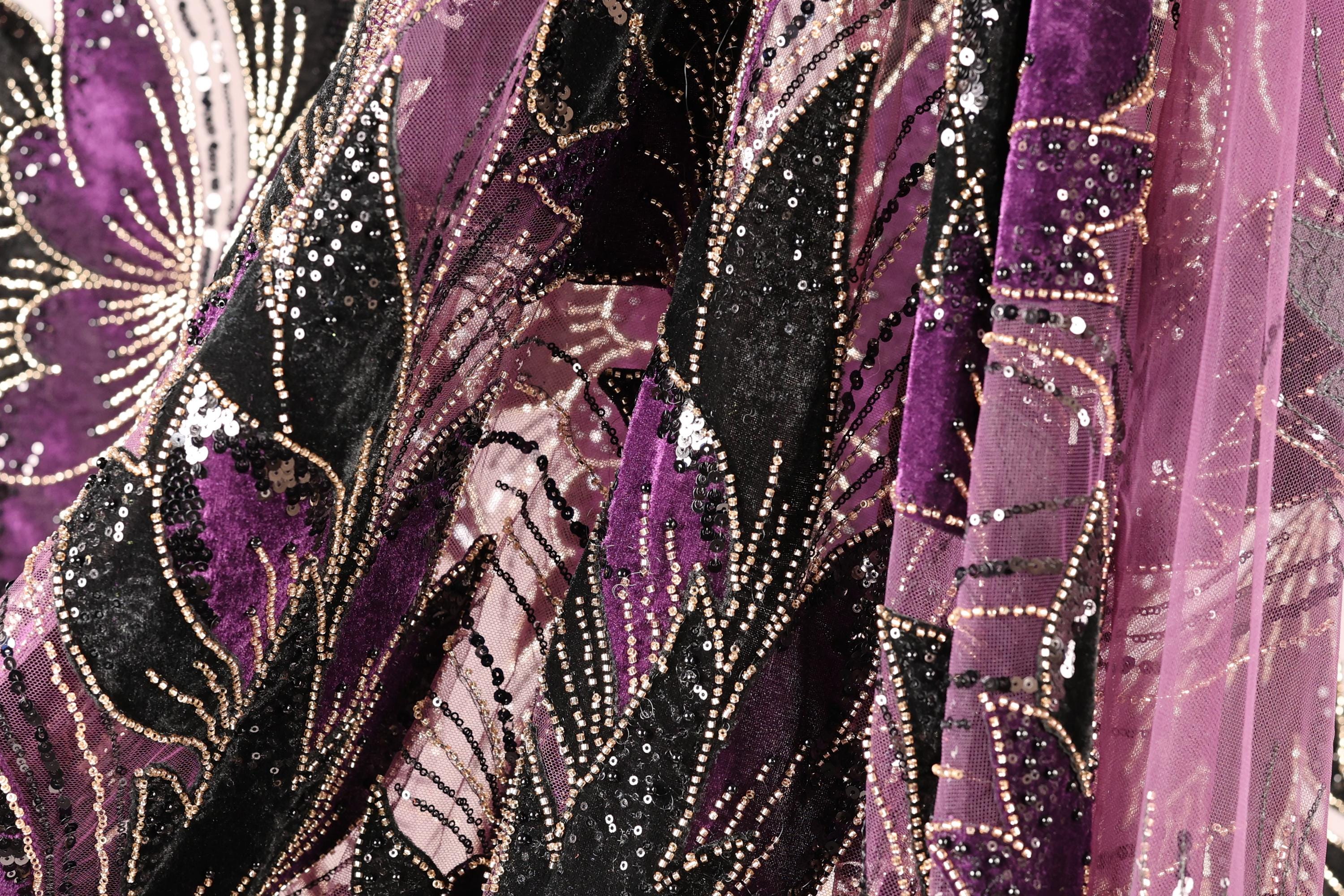 The image is a close-up of a fabric with a floral pattern, featuring a mix of purple and black colors. The fabric appears to be made of a shiny, sequined material, giving it a sparkling and luxurious appearance.