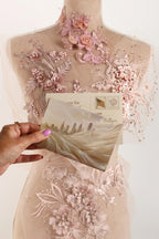A hand holding a postcard with a cityscape image, set against a background of a mannequin dress with intricate lace and floral embroidery.
