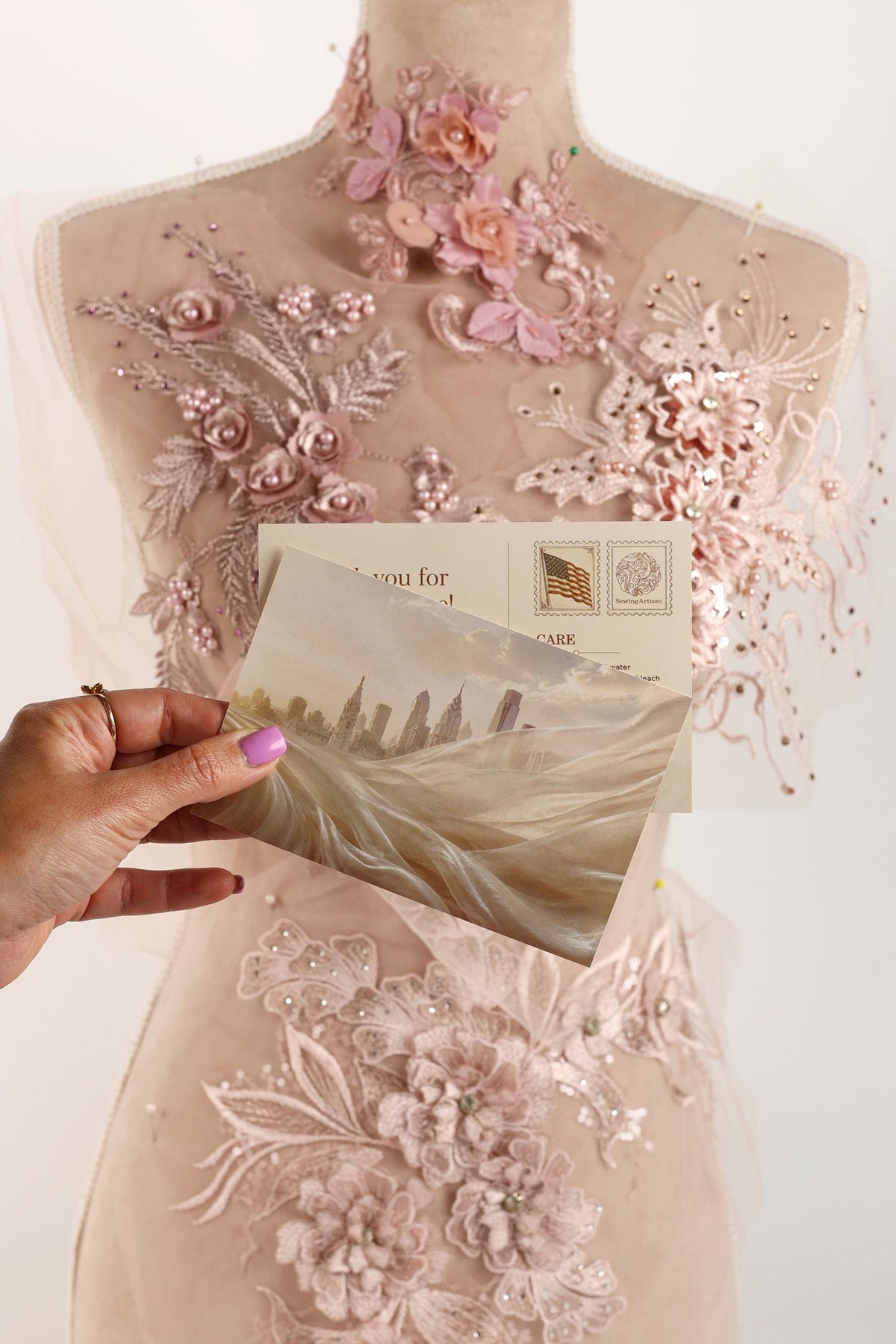 A hand holding a postcard with a cityscape image, set against a background of a mannequin dress with intricate lace and floral embroidery.