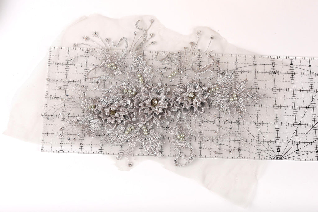 a decorative piece of fabric with a floral pattern, including a variety of flowers and leaves. The fabric appears to be a lace or sheer material, with a metallic or silver-colored appearance. The piece is placed on a transparent ruler, which is lying on a white surface.