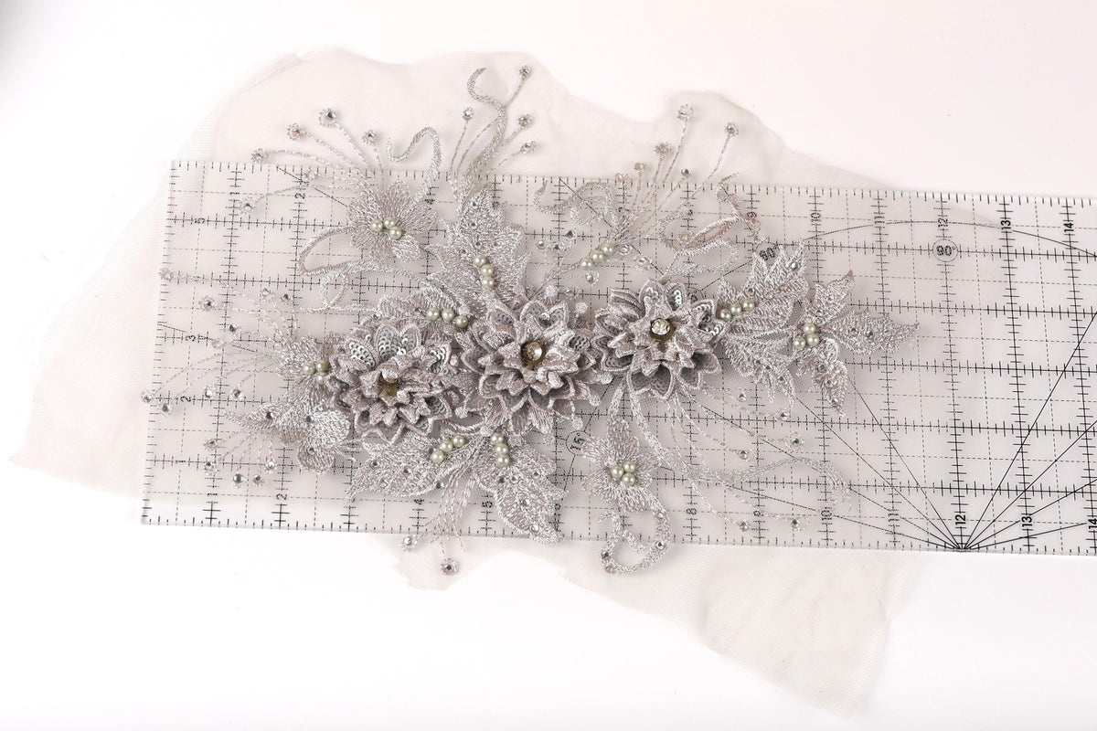 a decorative piece of fabric with a floral pattern, including a variety of flowers and leaves. The fabric appears to be a lace or sheer material, with a metallic or silver-colored appearance. The piece is placed on a transparent ruler, which is lying on a white surface.