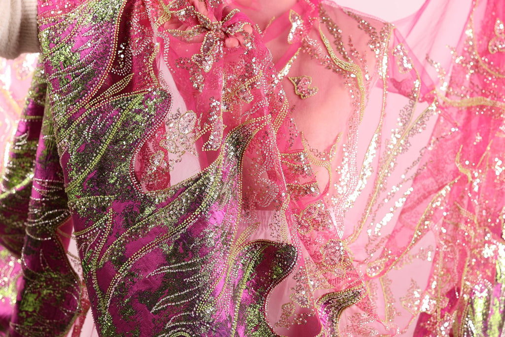 a close-up view of a vibrant, sequined dress with a pink and green color scheme. The dress appears to be made of sheer fabric, giving it a delicate and airy appearance. The intricate patterns and shimmering sequins create a visually striking and eye-catching design.