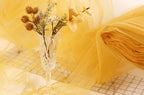 A clear glass vase filled with dried flowers and branches sits on a table covered with yellow fabric.
