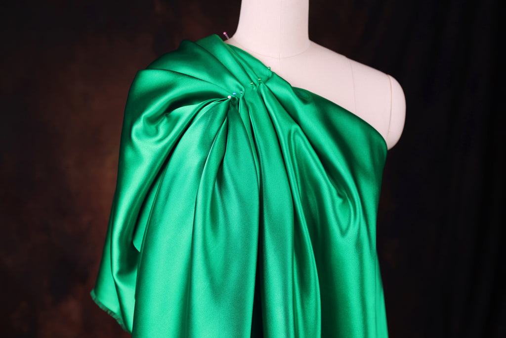 A mannequin wearing a green satin one-shoulder dress with a draped, flowing silhouette.