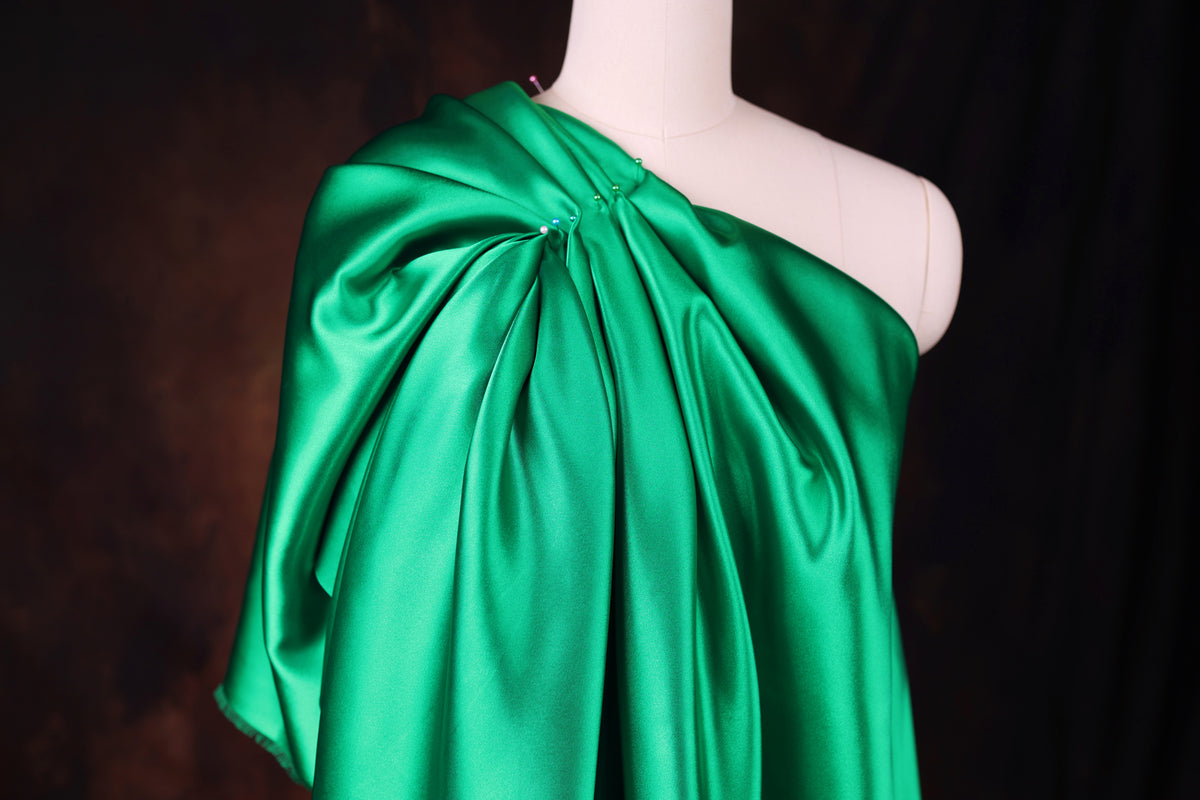 A mannequin wearing a green satin one-shoulder dress with a draped, flowing silhouette.