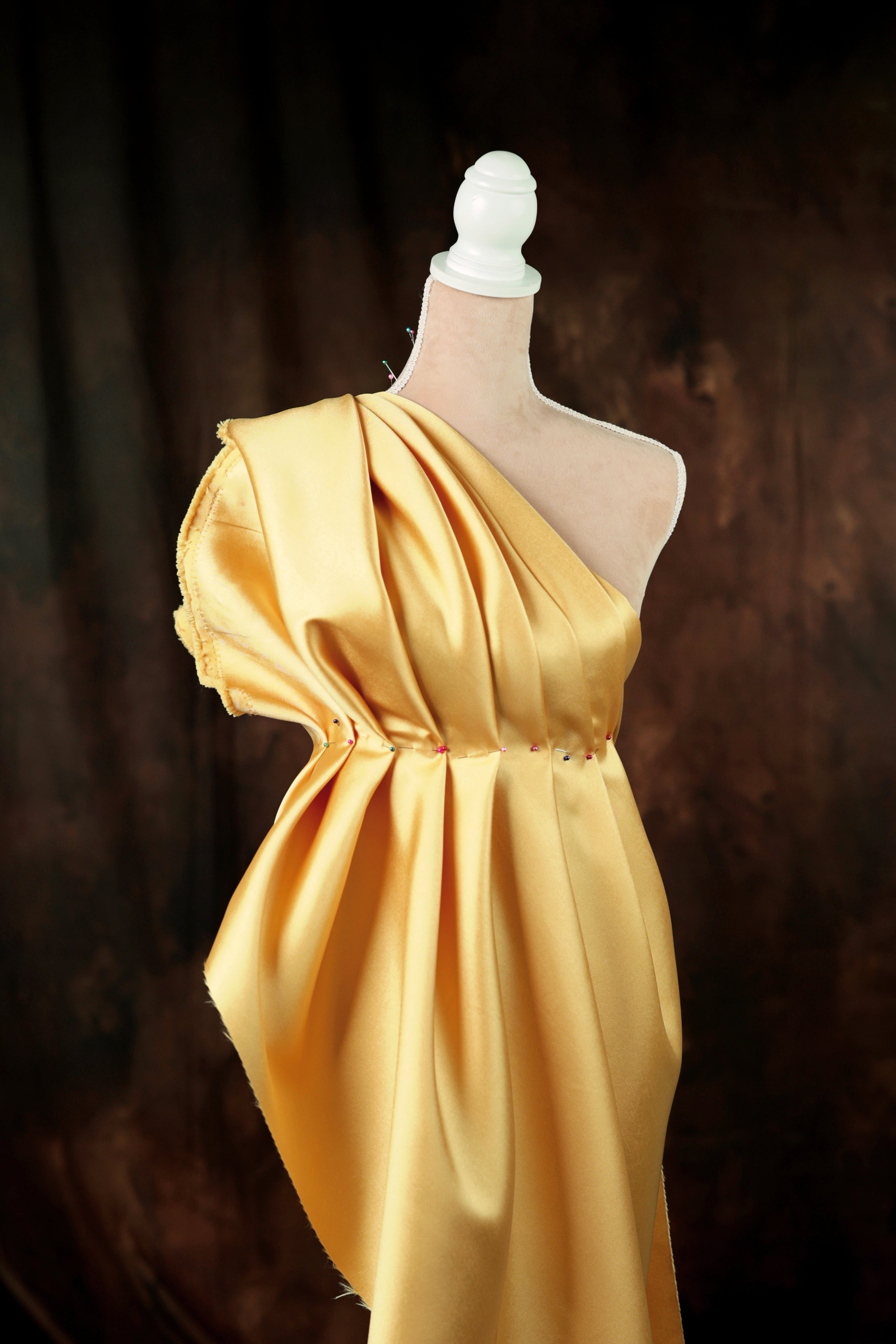 A mannequin wearing a gold, one-shoulder dress with a draped, flowing silhouette.