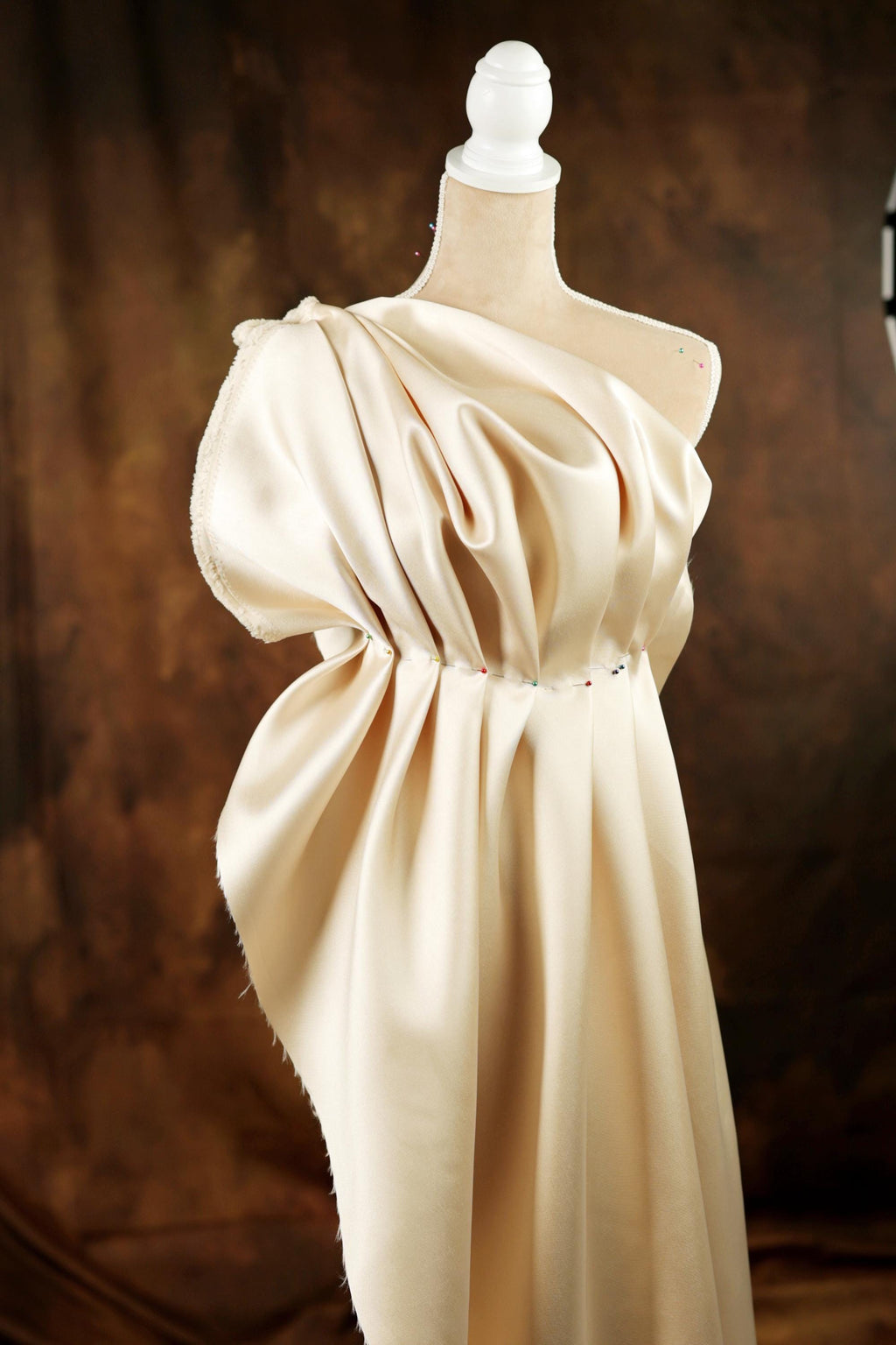 A mannequin wearing a beige, flowing dress with a draped, ruched neckline and a high collar.