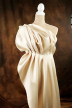 A mannequin wearing a beige, flowing dress with a draped, ruched neckline and a high collar.