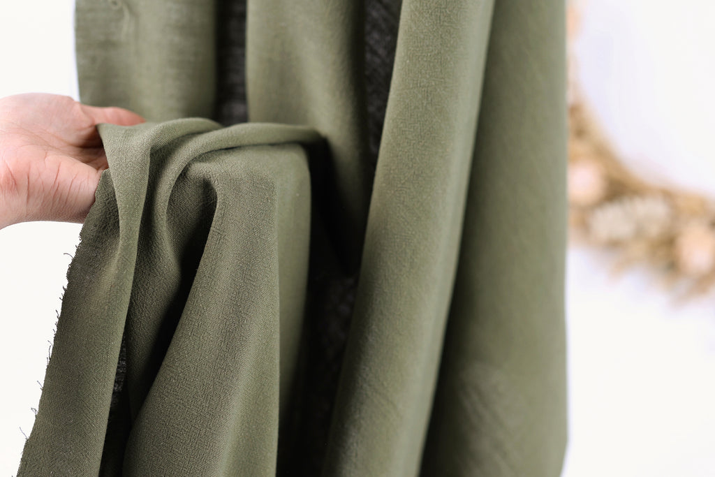 A hand is holding a piece of olive green fabric, which appears to be a type of cloth or textile.