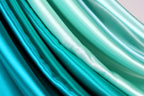 a close-up view of several pieces of fabric with a gradient of colors, including shades of blue, green, and white. The fabric appears to be made of silk or satin, and the colors are vibrant and rich, creating a visually striking and harmonious composition.