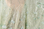 The image is a close-up view of a fabric with a floral pattern, featuring green leaves and sequins. The fabric appears to be a light green color, with a delicate and intricate design.