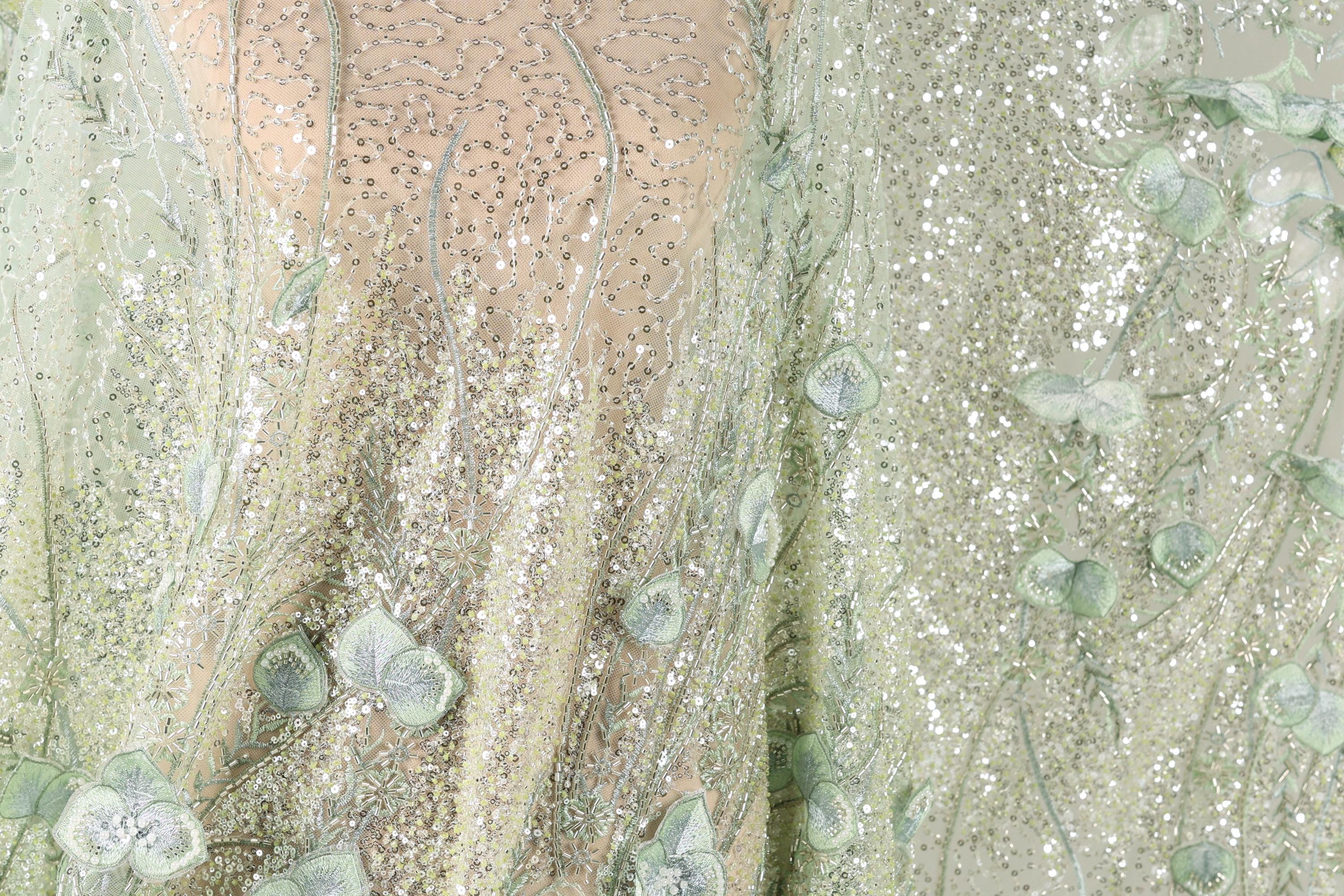 The image is a close-up view of a fabric with a floral pattern, featuring green leaves and sequins. The fabric appears to be a light green color, with a delicate and intricate design.