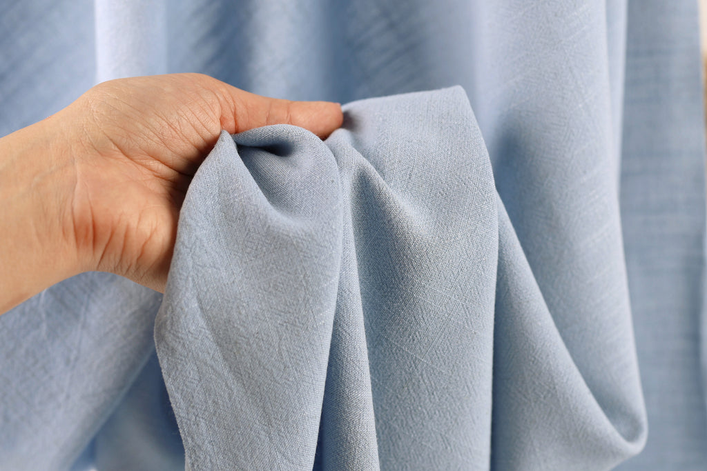 A hand is holding a piece of light blue fabric, possibly a curtain or textile, in front of a blurred background.