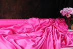 A close-up view of a crumpled, shiny pink satin fabric, with a vase of pink flowers visible in the background.