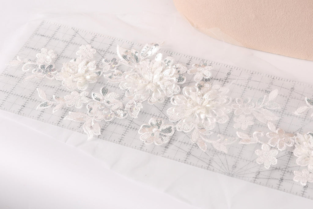a close-up view of a delicate, white lace fabric with intricate floral patterns. The fabric is spread out on a transparent surface, and a ruler is visible in the background, likely used for measuring or cutting the fabric.