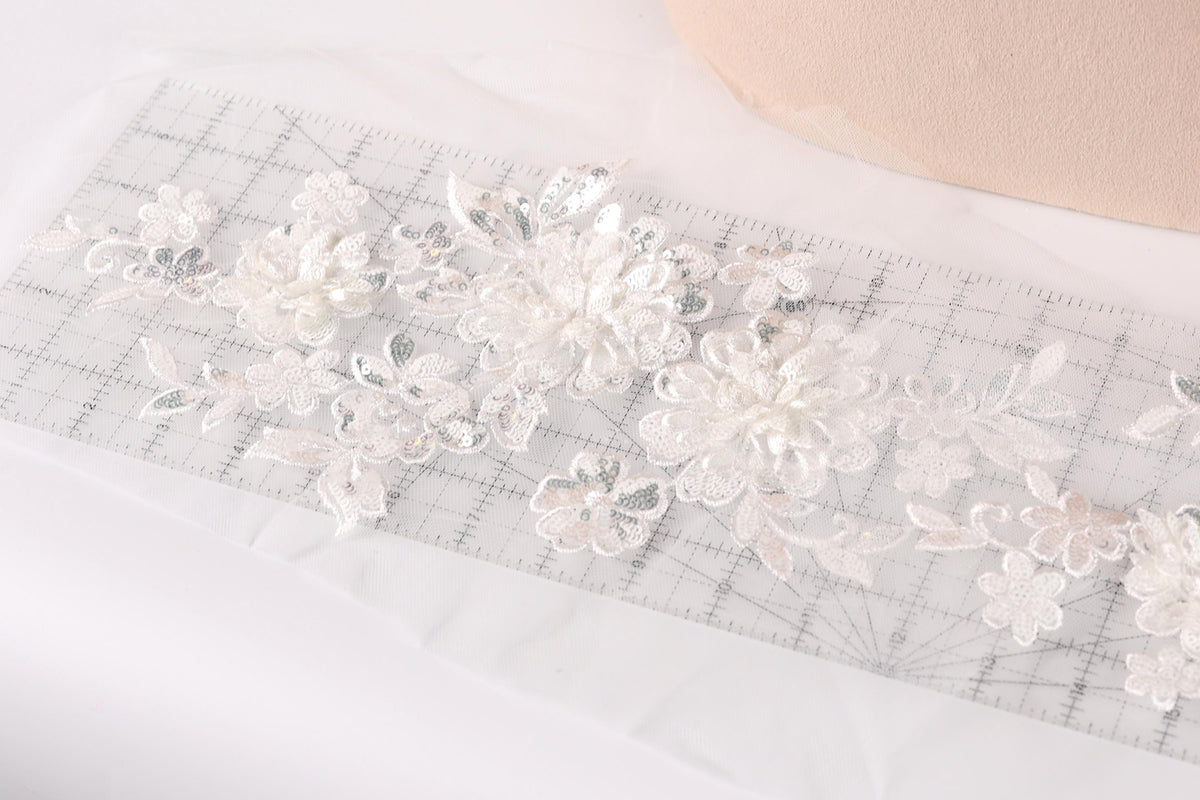 a close-up view of a delicate, white lace fabric with intricate floral patterns. The fabric is spread out on a transparent surface, and a ruler is visible in the background, likely used for measuring or cutting the fabric.