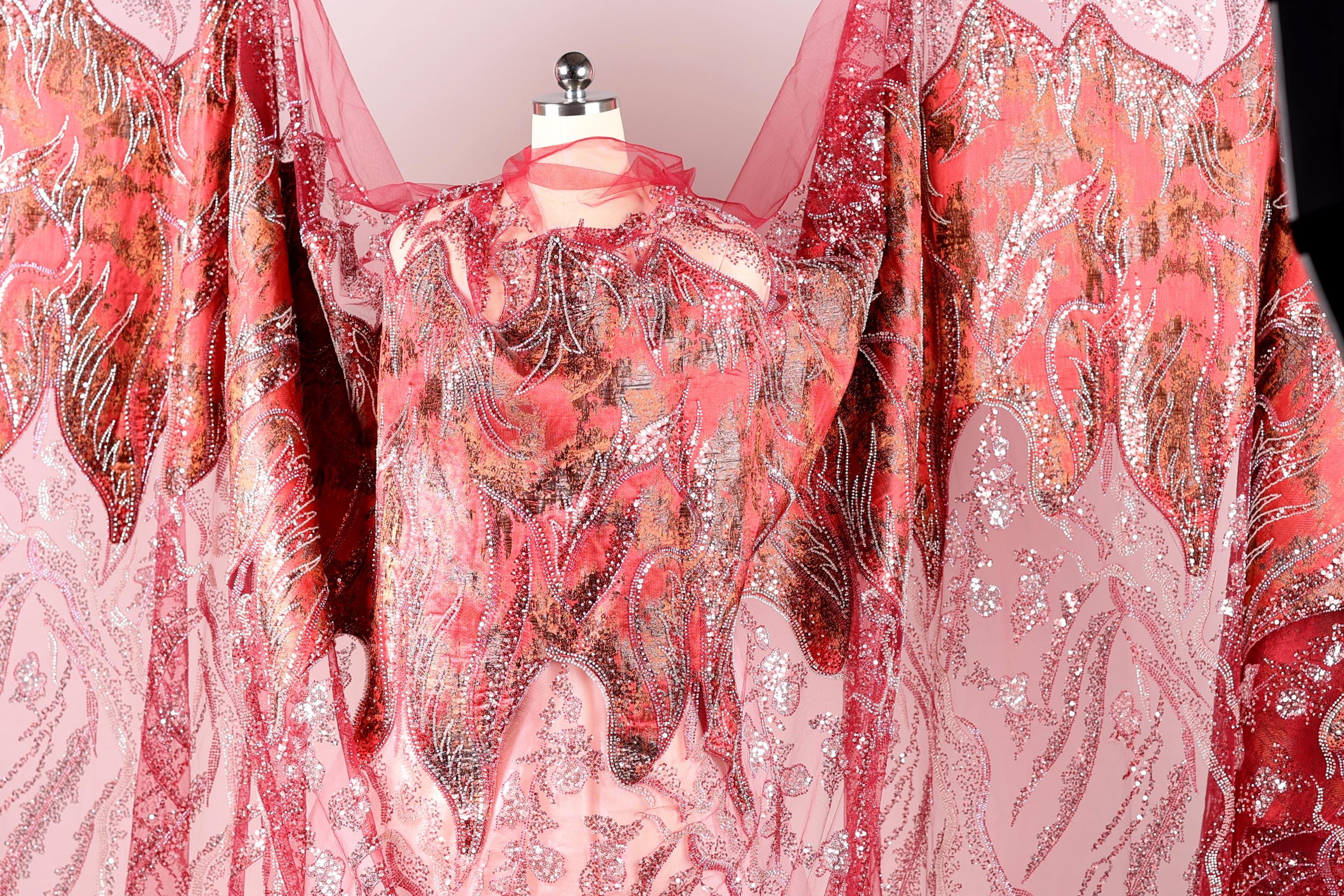 a vibrant, intricately designed dress with a striking pattern of red and pink hues. The dress appears to be made of a sheer fabric, giving it a delicate and airy appearance. The pattern on the dress is quite detailed, with a mix of floral and geometric elements that create a visually captivating design. The dress is displayed on a mannequin, showcasing its unique and eye-catching design.